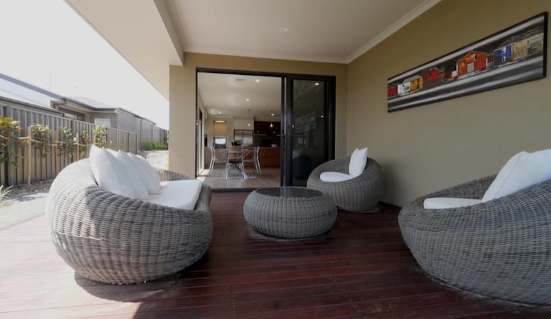 A rendered alfresco with timber decking and tub chairs looking back into the house